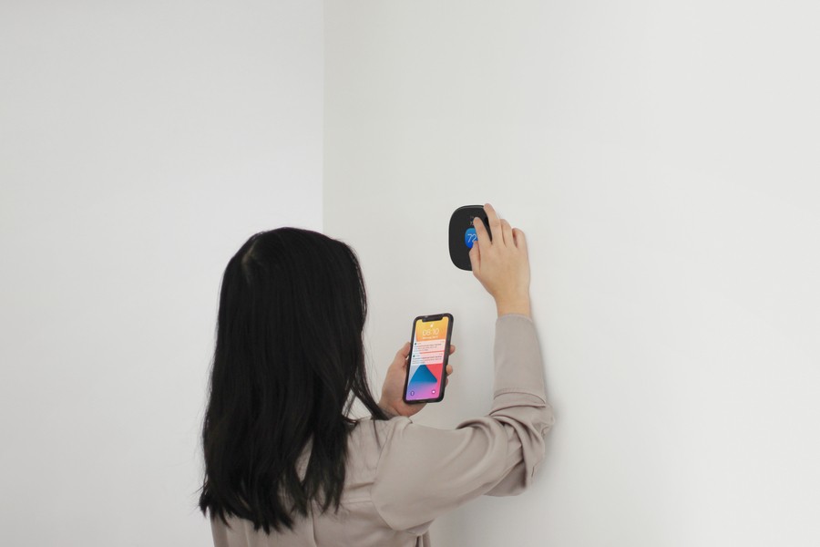 Person with long hair holding a smartphone while adjusting an Ecobee thermostat