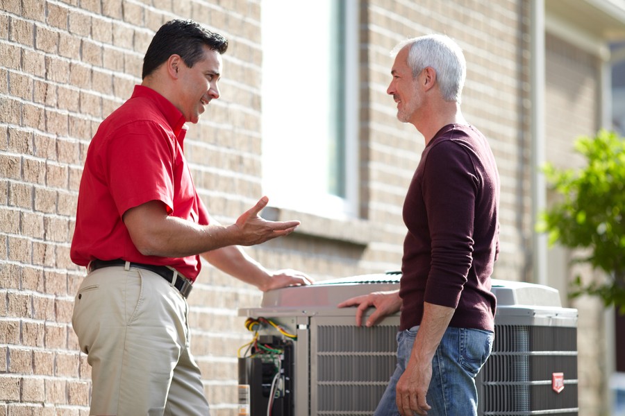 HVAC technician in red shirt explaining air conditioner maintenance to a smiling homeowner.