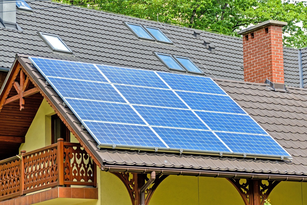 Mounted solar panels on a sloped residential roof with skylights and a brick chimney, surrounded by trees in a suburban setting.