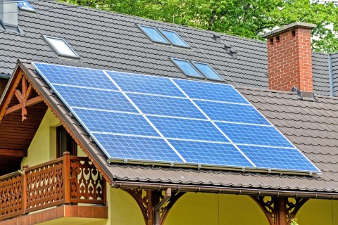 Mounted solar panels on a sloped residential roof with skylights and a brick chimney, surrounded by trees in a suburban setting.