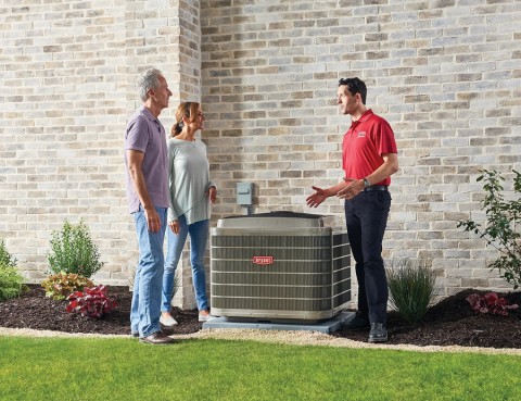 HVAC technician in a red polo shirt explaining a newly installed Bryant air conditioning unit to a smiling couple outside their home, next to a brick wall and landscaped yard.