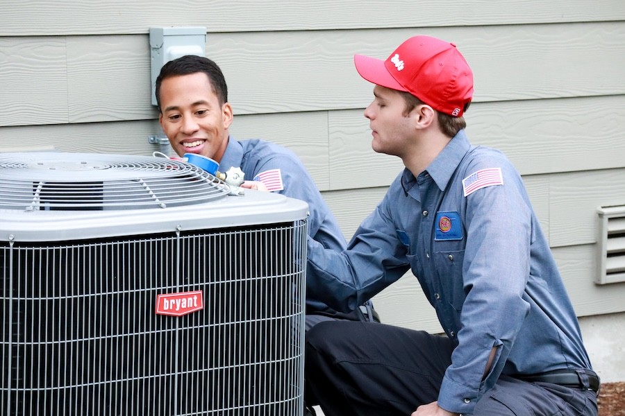 Two Bob’s Heating technicians crouch near an outdoor Bryant unit to perform service work.