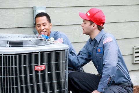 Two Bob’s Heating technicians crouch near an outdoor Bryant unit to perform service work.